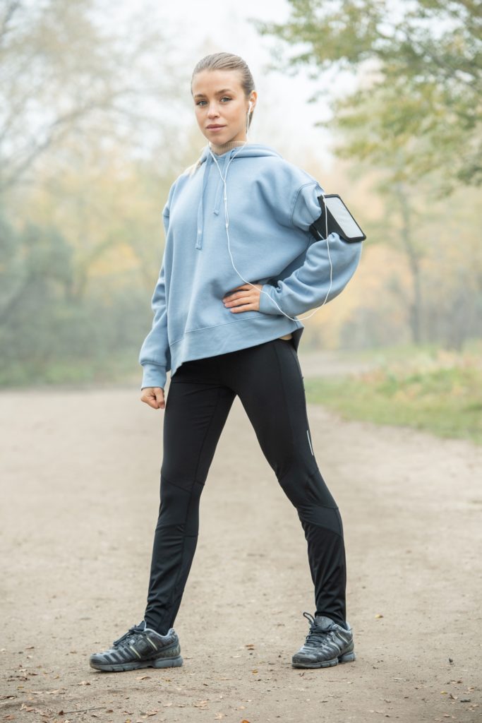 Young serious sportswoman in leggins and hoodie listening to music in earphones